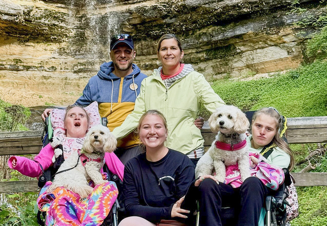 Paul and Kathy with another woman, along with their daughters Katelyn and Madelyn, each holding a dog on their laps, with a waterfall behind them.