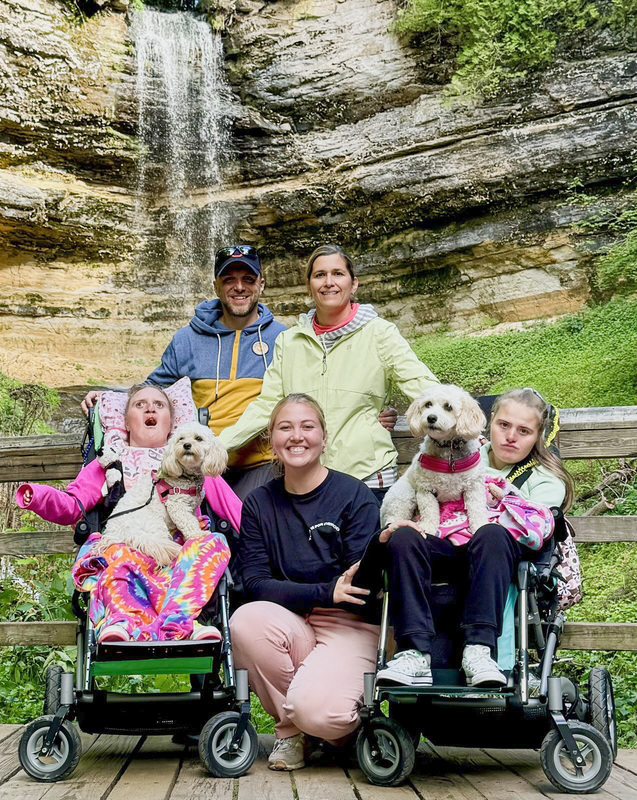 Paul and Kathy with another woman, along with their daughters Katelyn and Madelyn, each holding a dog on their laps, with a waterfall behind them.