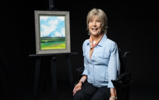 Joni Eareckson Tada smiling and posing next to her artwork titled "Kathren's Cloud," which features patches of flowers and two mountains beneath bright clouds and a blue sky.