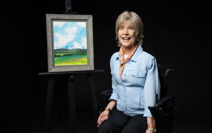 Joni Eareckson Tada smiling and posing next to her artwork titled "Kathren's Cloud," which features patches of flowers and two mountains beneath bright clouds and a blue sky.