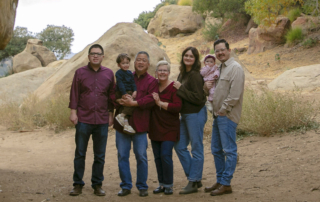 Chris Sue carrying his grandson, with Kendall, Stephanie, his daughter-in-law, and his eldest son holding his baby, all posing for the camera outdoors.