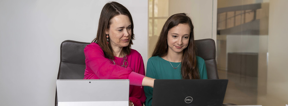 Two women sitting side by side, looking at a laptop screen with focused expressions.