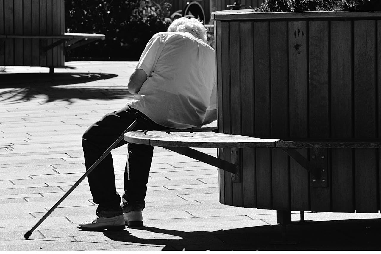 An elderly man sitting alone on a wooden chair outdoors with a cane resting beside him