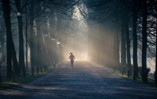 A woman running on a rough road lined with trees, with sunlight gleaming through the dark foliage.