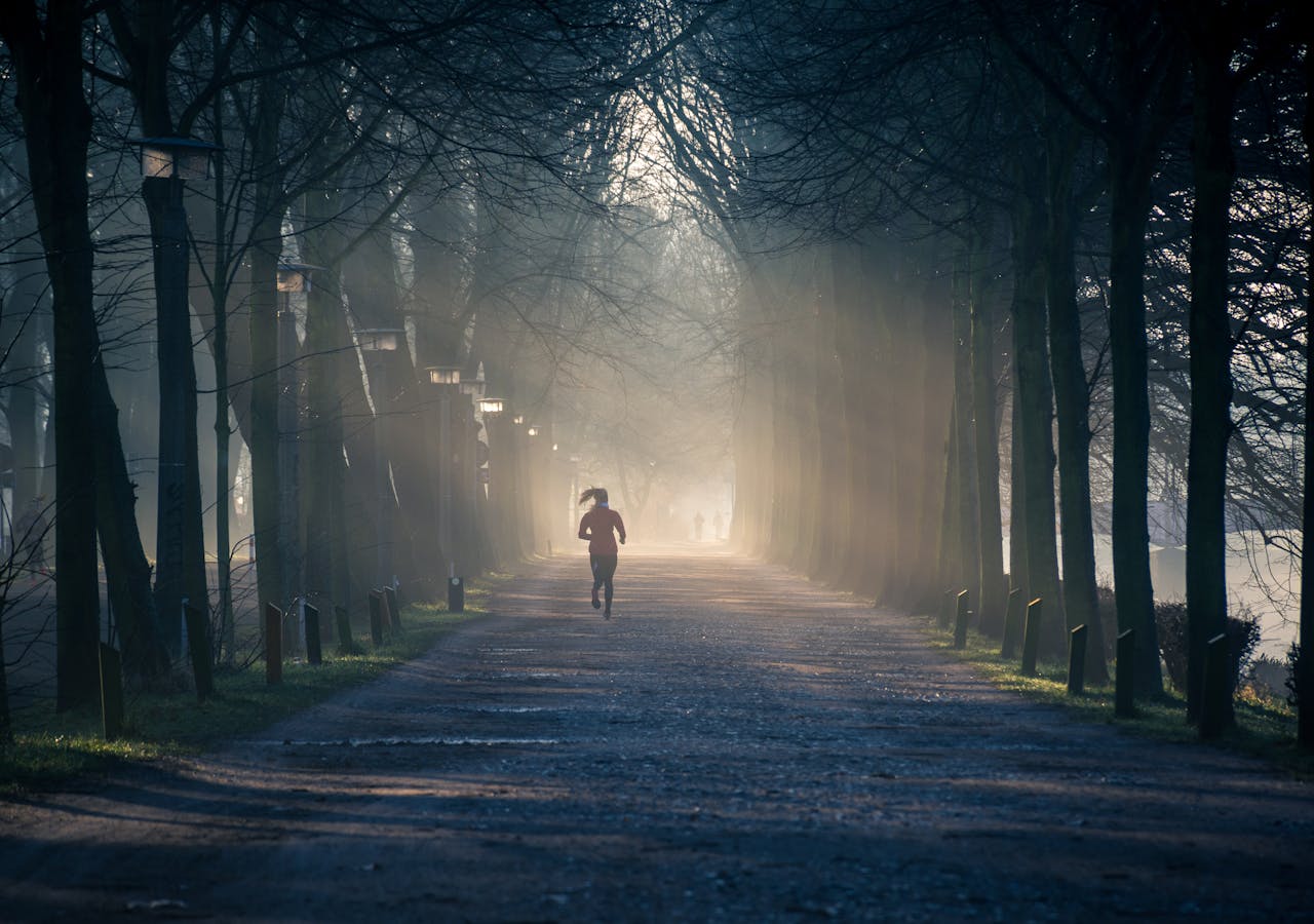 A woman running on a rough road lined with trees, with sunlight gleaming through the dark foliage.