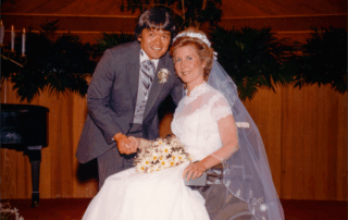 A picture of Joni and Ken on their wedding day at the altar. Joni is seated in her wheelchair with her wedding dress on, Ken leaning over her. Both smiling at the camera.