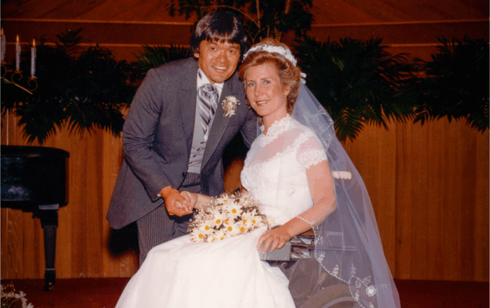 A picture of Joni and Ken on their wedding day at the altar. Joni is seated in her wheelchair with her wedding dress on, Ken leaning over her. Both smiling at the camera.