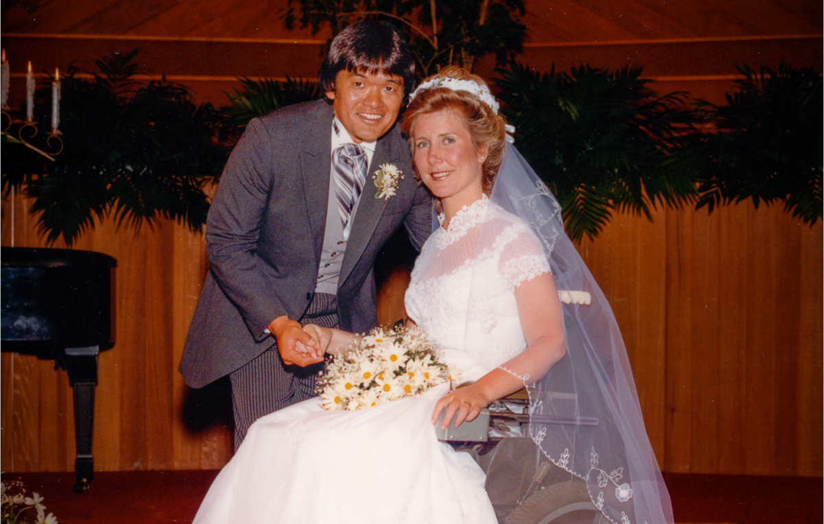 A picture of Joni and Ken on their wedding day at the altar. Joni is seated in her wheelchair with her wedding dress on, Ken leaning over her. Both smiling at the camera.