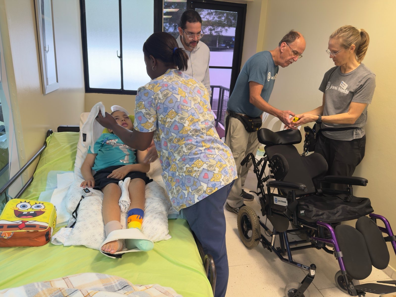 Carlito lying on a green bed while people in Joni and Friends shirts measure a wheelchair beside him, preparing for a custom fit.