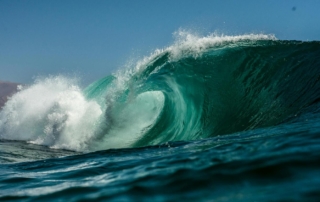 A powerful turquoise wave curls and crashes in the ocean under a clear blue sky, with mist spraying from the impact and distant mountains faintly visible in the background.
