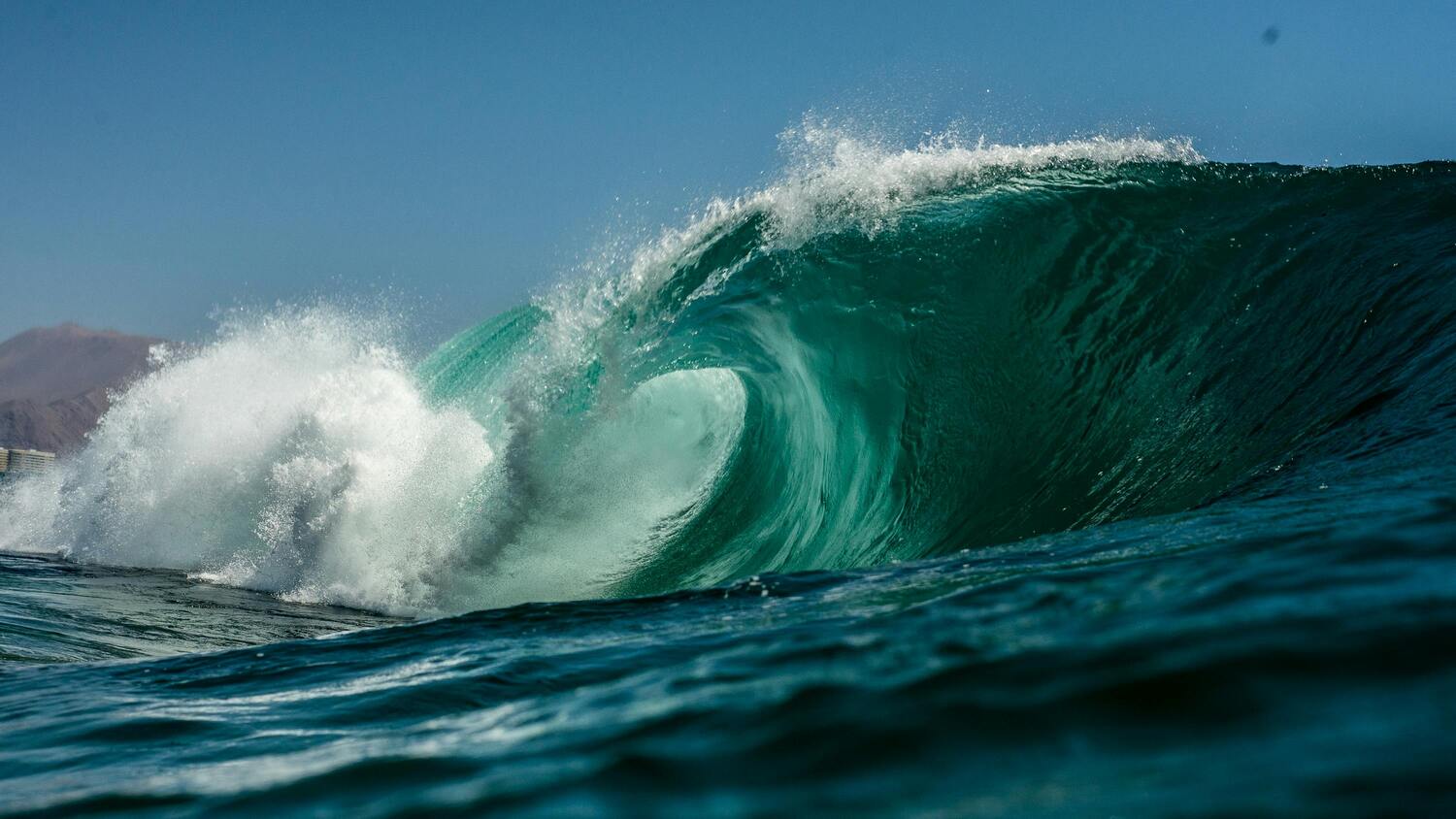 A powerful turquoise wave curls and crashes in the ocean under a clear blue sky, with mist spraying from the impact and distant mountains faintly visible in the background.