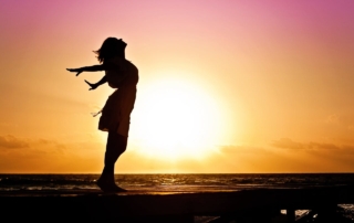 Silhouette of a woman standing by the ocean, feeling the breeze as the sun sets in the background.
