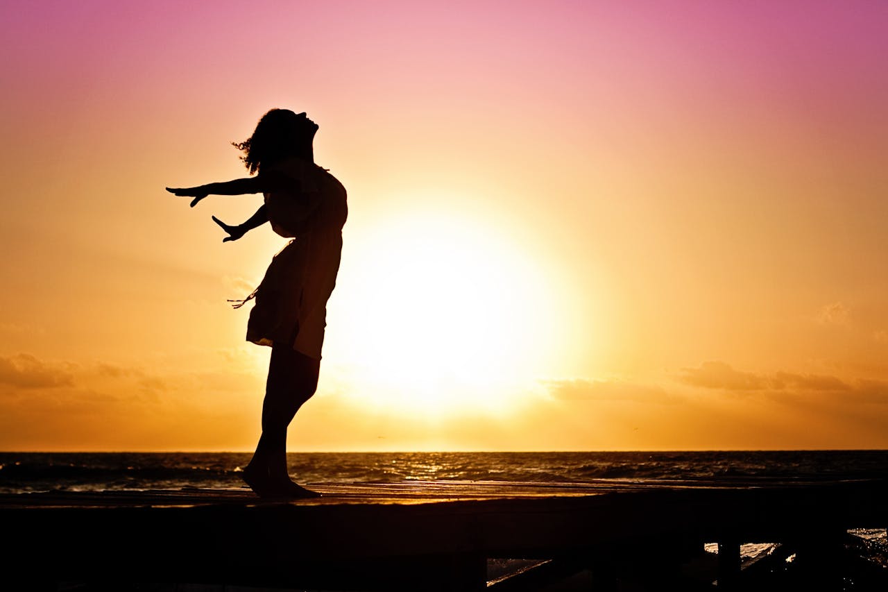 Silhouette of a woman standing by the ocean, feeling the breeze as the sun sets in the background.
