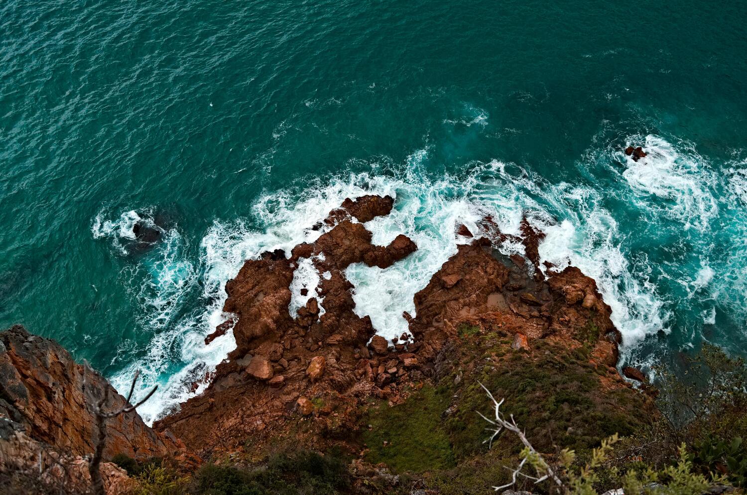 A top-down view of a blue ocean crashing against rugged brown rocks.