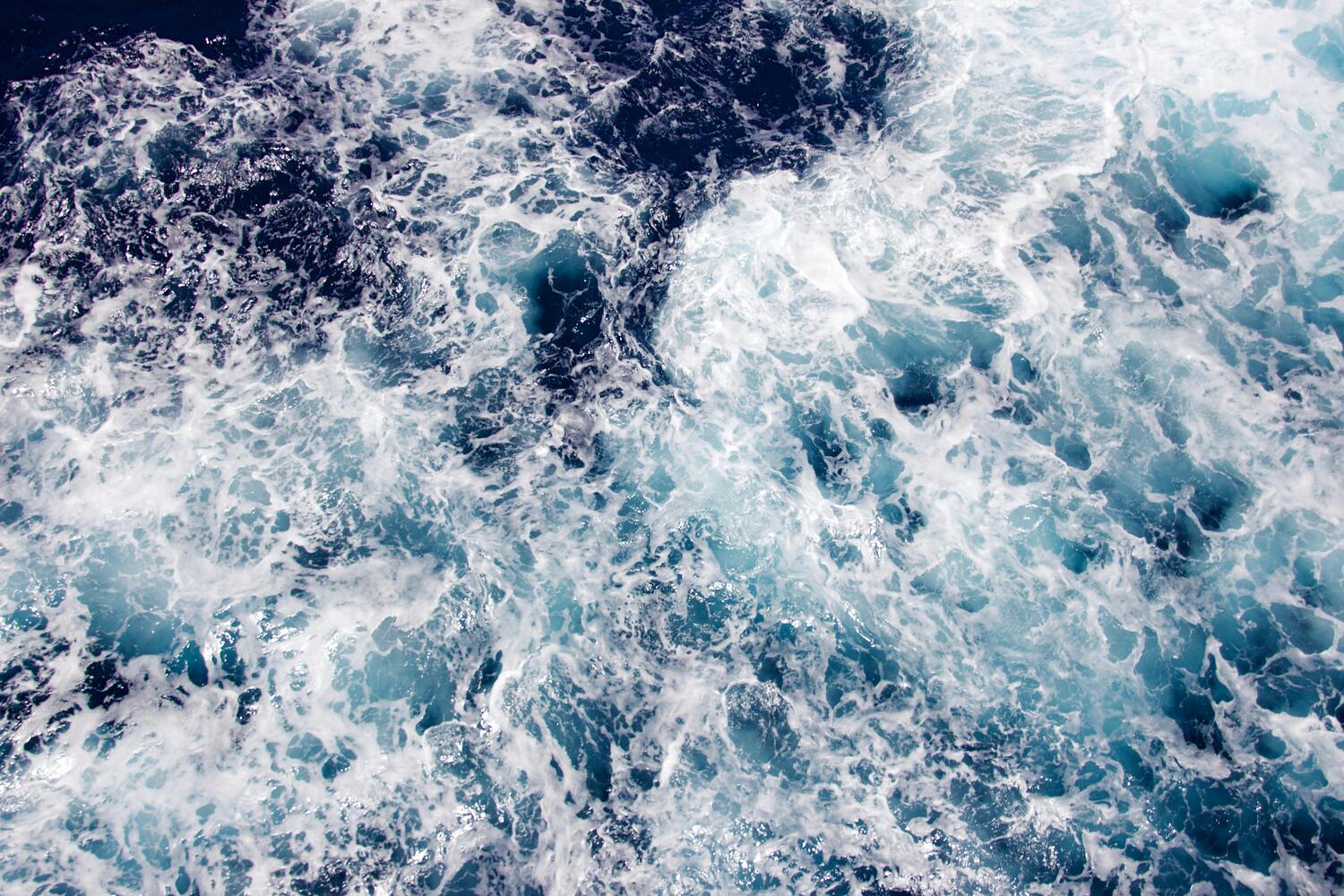 A top-down view of ocean water with white foam and swirling shades of deep blue and turquoise.