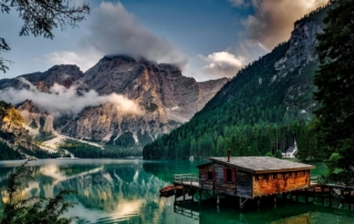 A serene mountain lake surrounded by pine-covered slopes and towering rocky peaks, with low clouds drifting near the summits. A rustic wooden cabin on stilts sits over the water in the foreground, with a small dock and boats nearby.