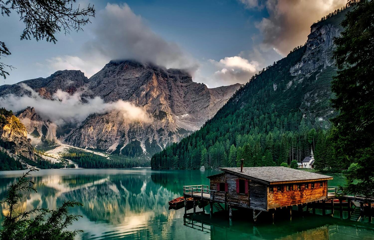 A serene mountain lake surrounded by pine-covered slopes and towering rocky peaks, with low clouds drifting near the summits. A rustic wooden cabin on stilts sits over the water in the foreground, with a small dock and boats nearby.