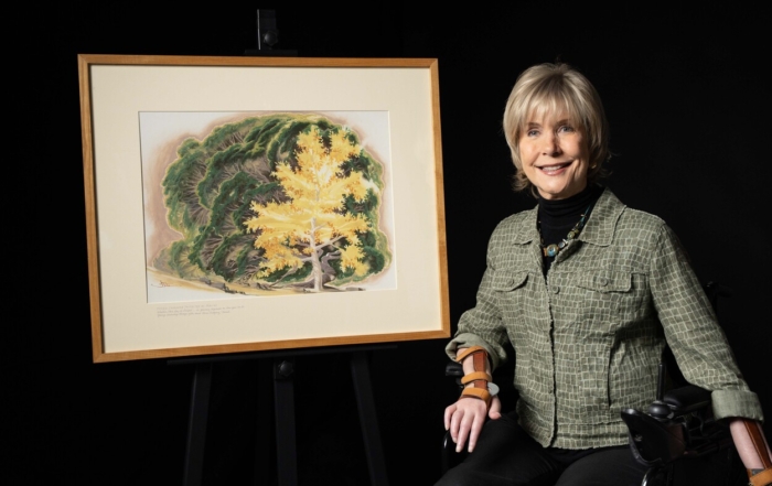 A woman in a wheelchair, identified as Joni Eareckson Tada, smiles beside one of her framed paintings on an easel. The artwork shows a single, youthful tree standing tall in front of a dense grouping of older, rounded bushes.