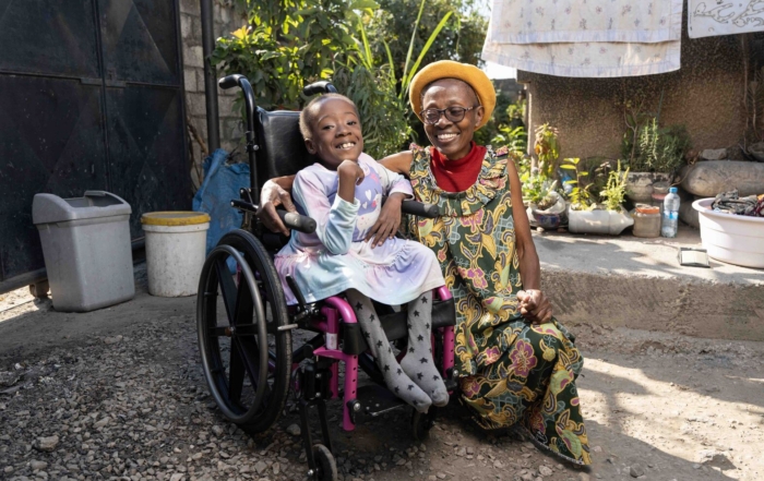 Diana, sitting in her new wheelchair, smiles excitedly at the camera while her aunt sits beside her, smiling warmly at Diana.