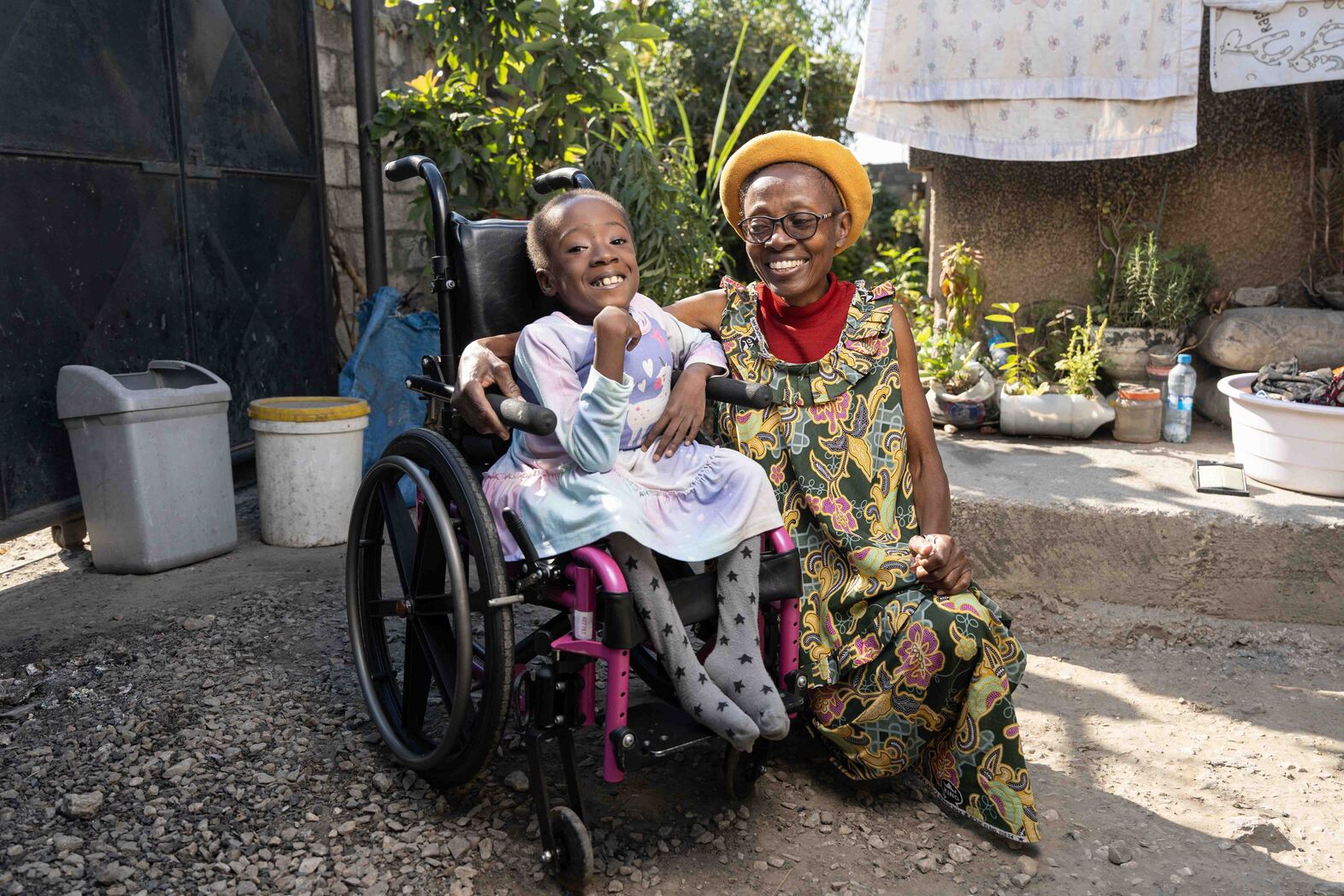Diana, sitting in her new wheelchair, smiles excitedly at the camera while her aunt sits beside her, smiling warmly at Diana.