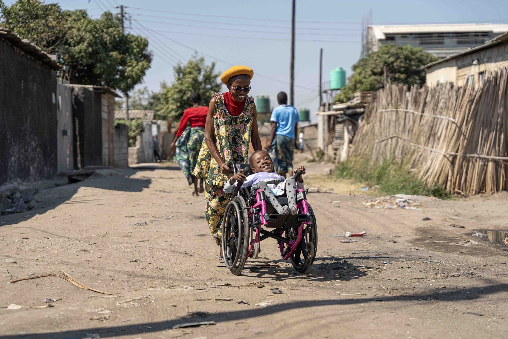 Diana sits in her wheelchair, smiling, while her aunt pushes her from behind, also smiling.