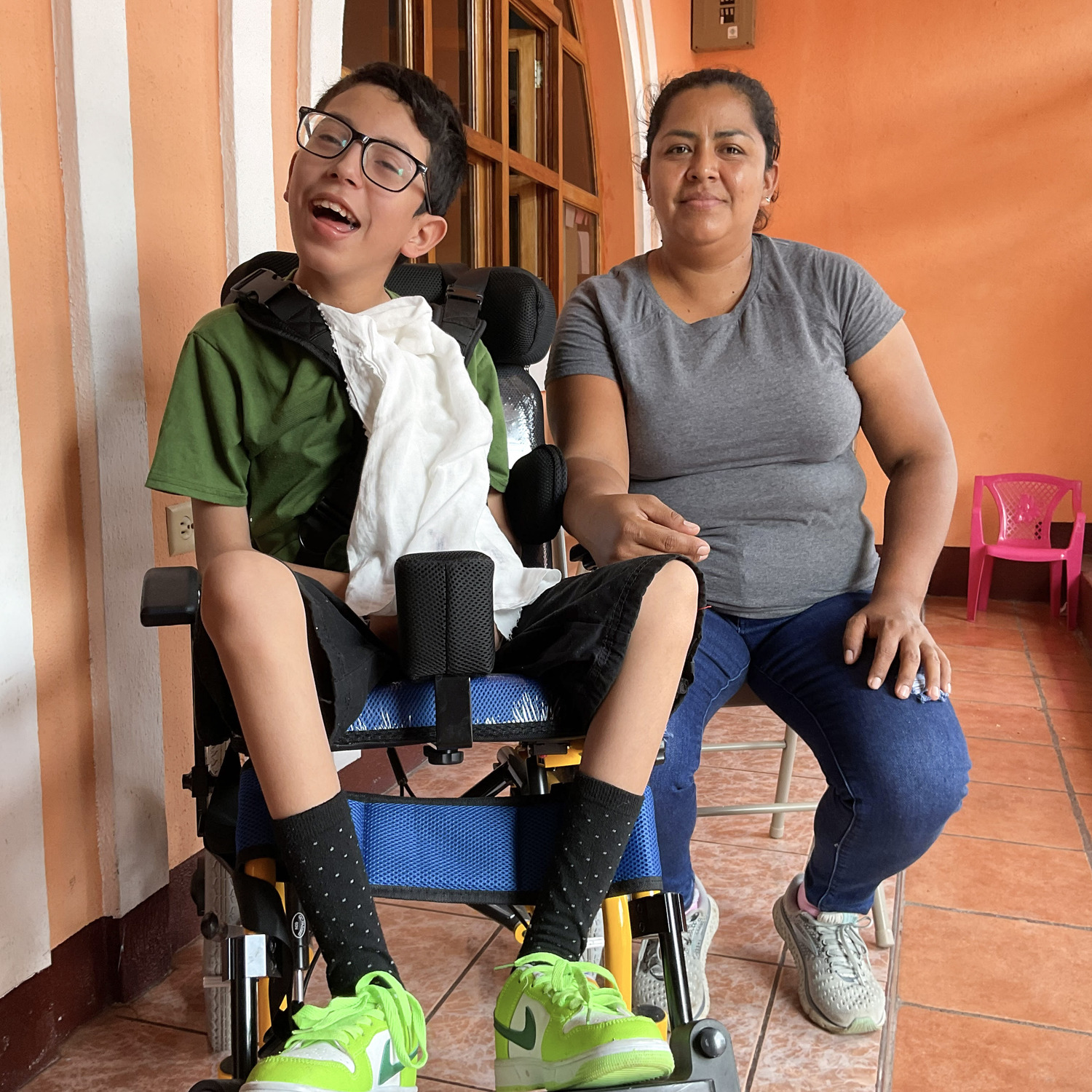 Axel Rios, a smiling young boy in a wheelchair, sits on the left wearing glasses and a bib. His mother sits beside him on the right, resting her hand on his chair. They are indoors on a tiled floor with walls and a window in the background.