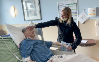 Jason Kolb in a hospital bed, his wife gently holding his forehead with one hand and his hand with the other.