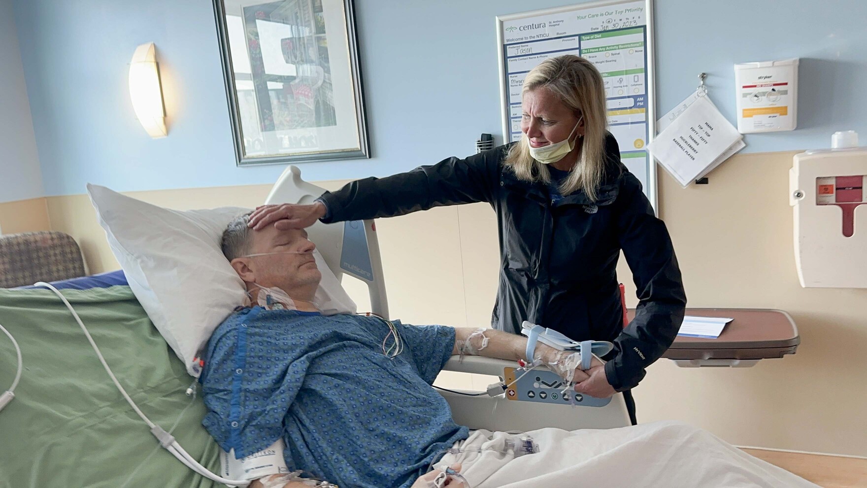 Jason Kolb in a hospital bed, his wife gently holding his forehead with one hand and his hand with the other.