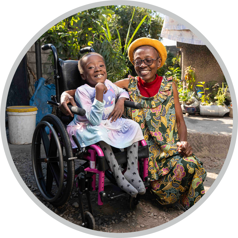 Diana, sitting in her new wheelchair, smiles excitedly at the camera while her aunt sits beside her, smiling warmly at Chichi.
