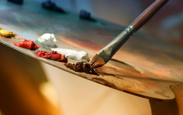 A close-up of a wooden painter’s palette with dabs of oil paint in red, yellow, white, and brown. A paintbrush is seen picking up brown paint from the palette.
