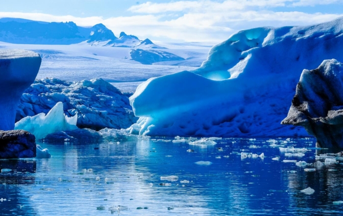 A stunning icy landscape featuring large blue glaciers and floating chunks of ice in a cold, reflective body of water, with snow-covered mountains visible in the background under a partly cloudy sky.