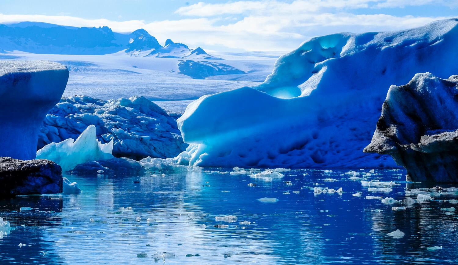 A stunning icy landscape featuring large blue glaciers and floating chunks of ice in a cold, reflective body of water, with snow-covered mountains visible in the background under a partly cloudy sky.