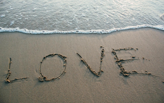 The word “LOVE” written in the sand near the shoreline, with gentle ocean waves approaching the letters.