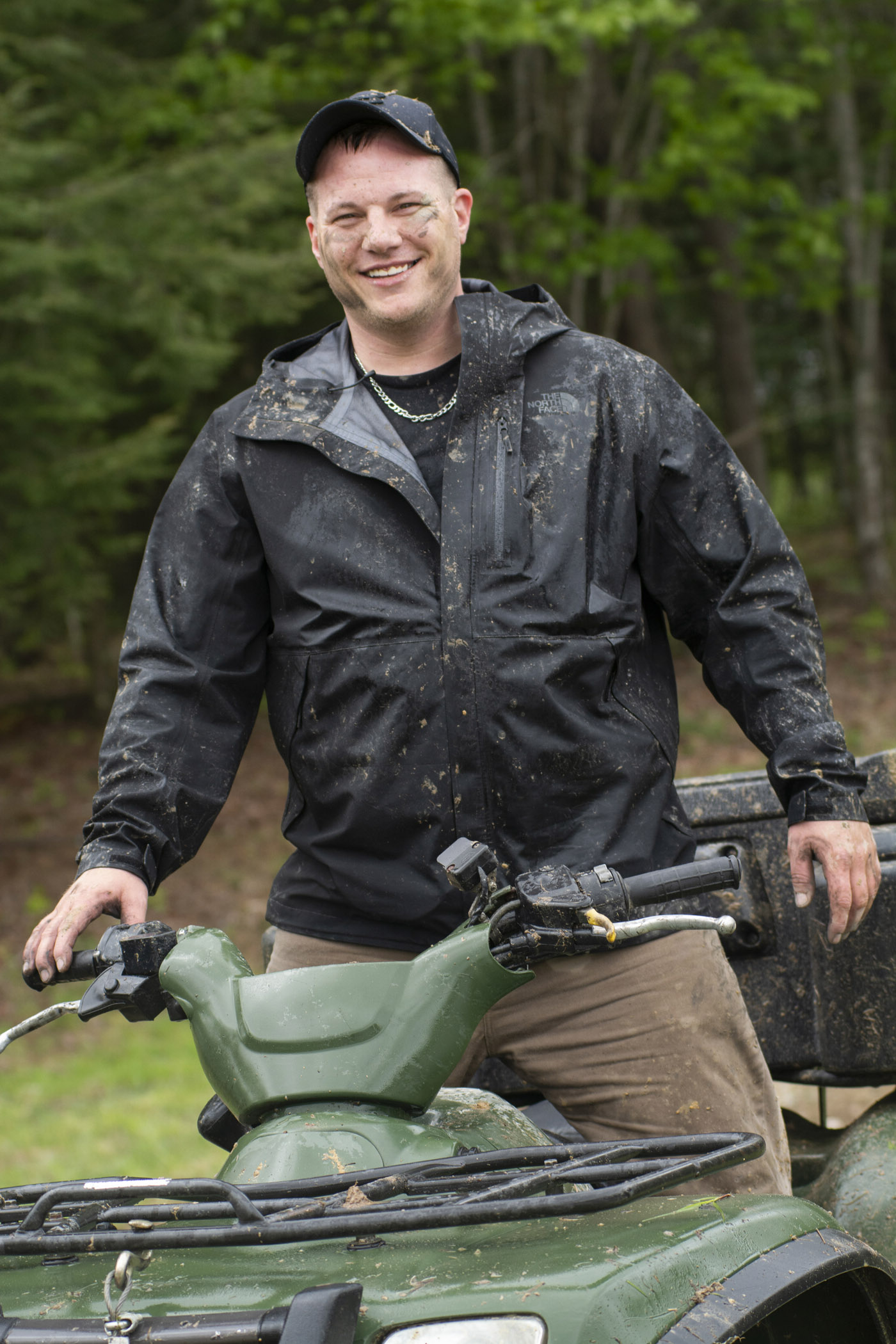 Mark Stallion smiles happily for the camera, his face and body covered in mud as he rides an all-terrain vehicle.