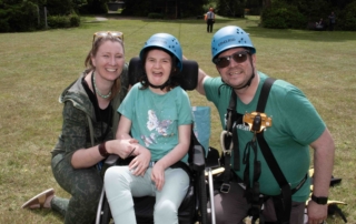 Sparrow in her wheelchair, wearing a helmet and smiling widely, with Lauren and Jon on either side of her.