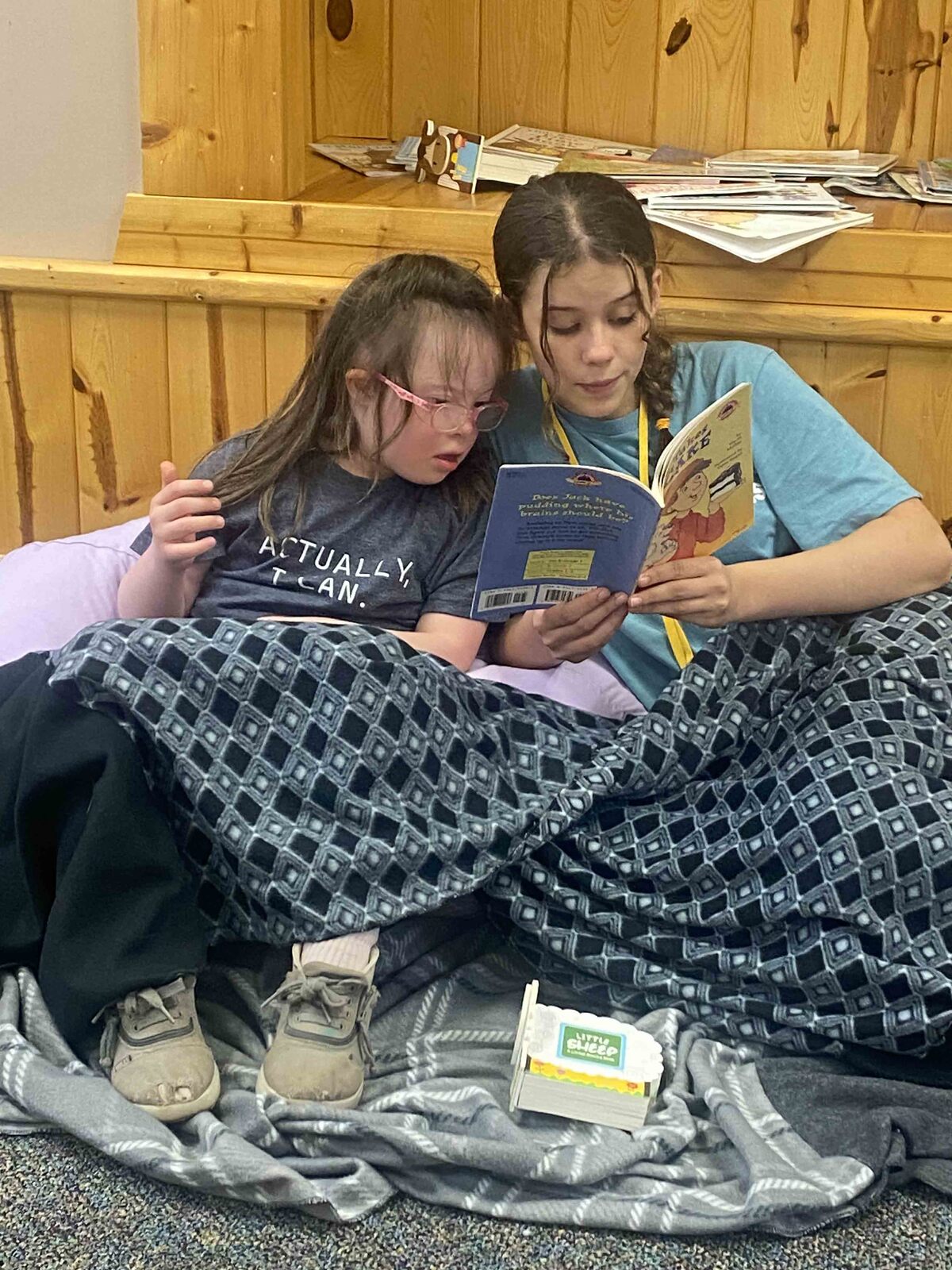 Elizabeth Magee sits on the left, wearing glasses and a shirt that says “ACTUALLY I CAN,” closely reading a book with another girl beside her. They are wrapped in a patterned blanket and sitting on the floor in front of a wooden shelf filled with books.