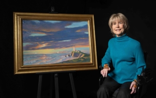 Joni Eareckson Tada smiling and posing next to her artwork titled "The Lighthouse". The painting depicts a lighthouse on a cliff overlooking the ocean at sunset, with waves crashing against the shore and sailboats in the distance.