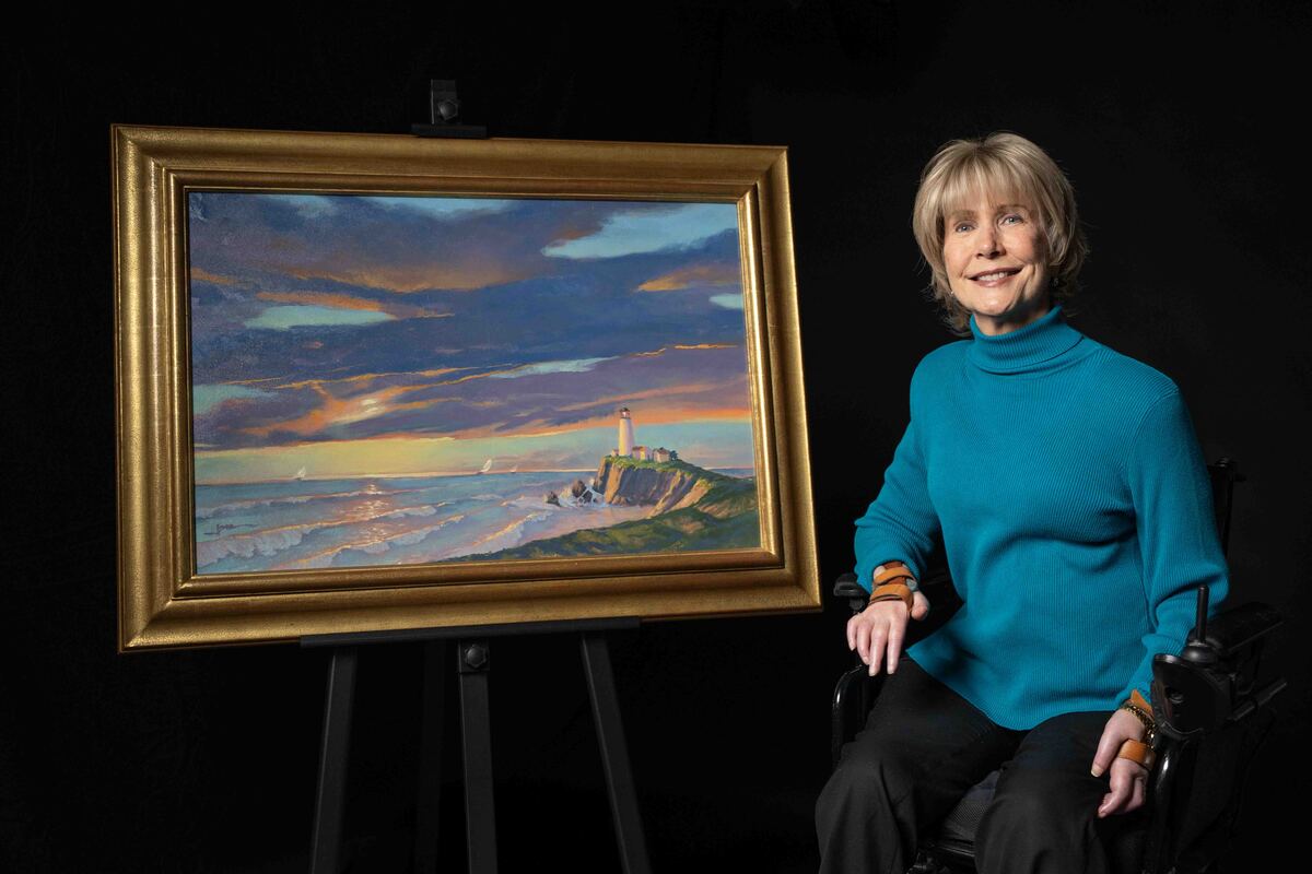 Joni Eareckson Tada smiling and posing next to her artwork titled "The Lighthouse". The painting depicts a lighthouse on a cliff overlooking the ocean at sunset, with waves crashing against the shore and sailboats in the distance.