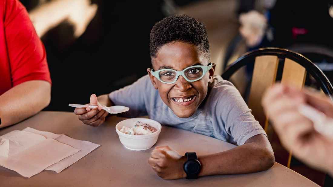 A boy with blue glasses stops eating ice cream to smile for a photo