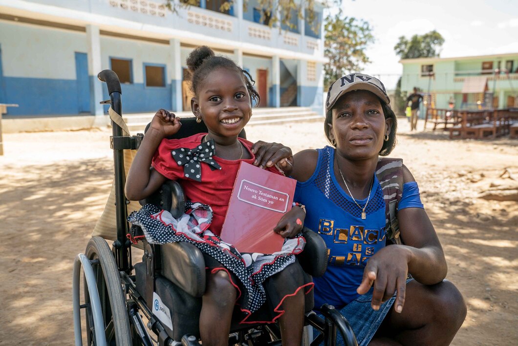 A joyful little girl in a wheelchair holding a book titled 'Nouvo Testaman an ak Som Yo' (The New Testament and Psalms), raising one arm in excitement, with a woman beside her.