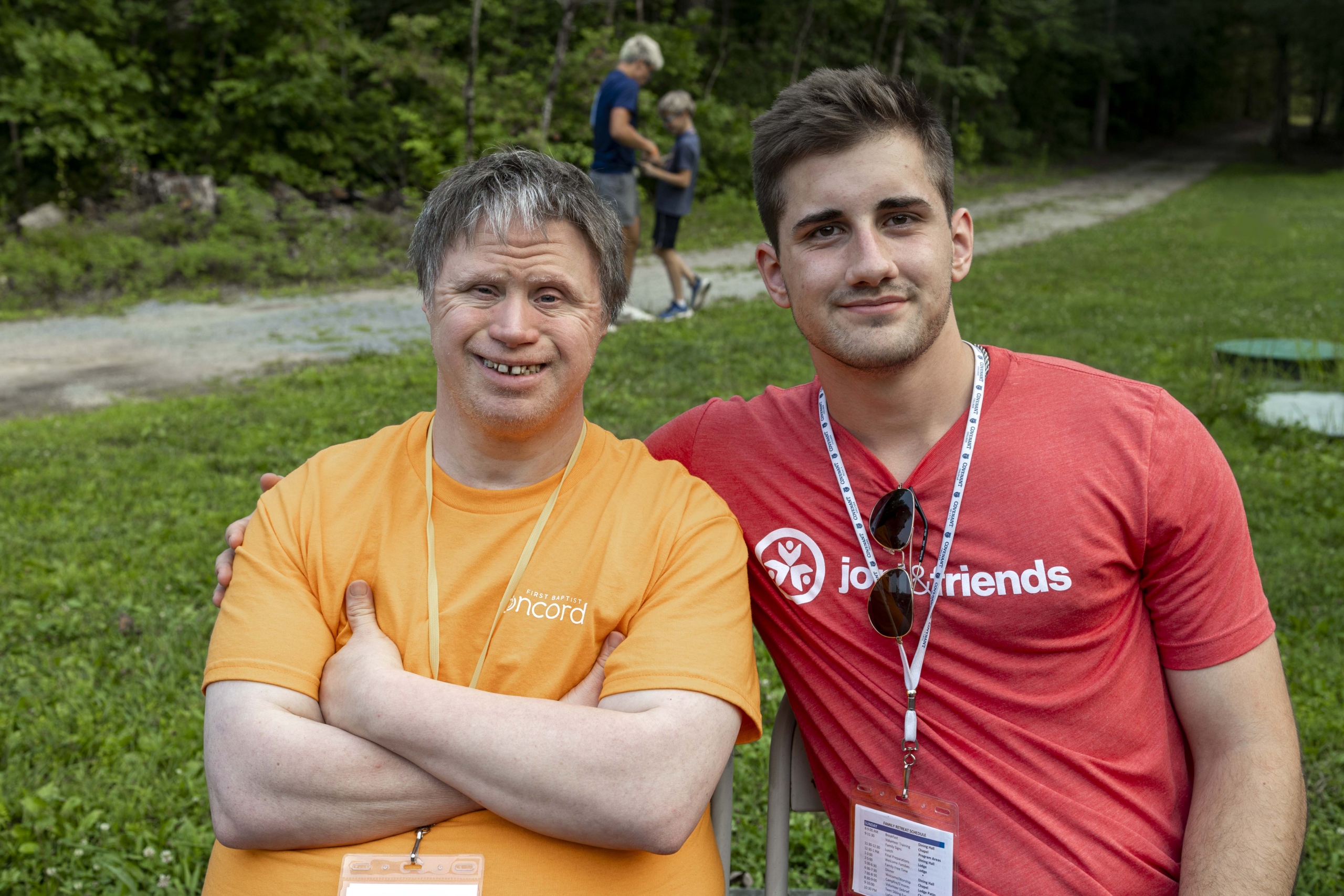 A man with Down syndrome and his buddy smiling and posing for the camera during a Family Retreat at Joni and Friends, in an outdoor setting with green grass and trees in the background.