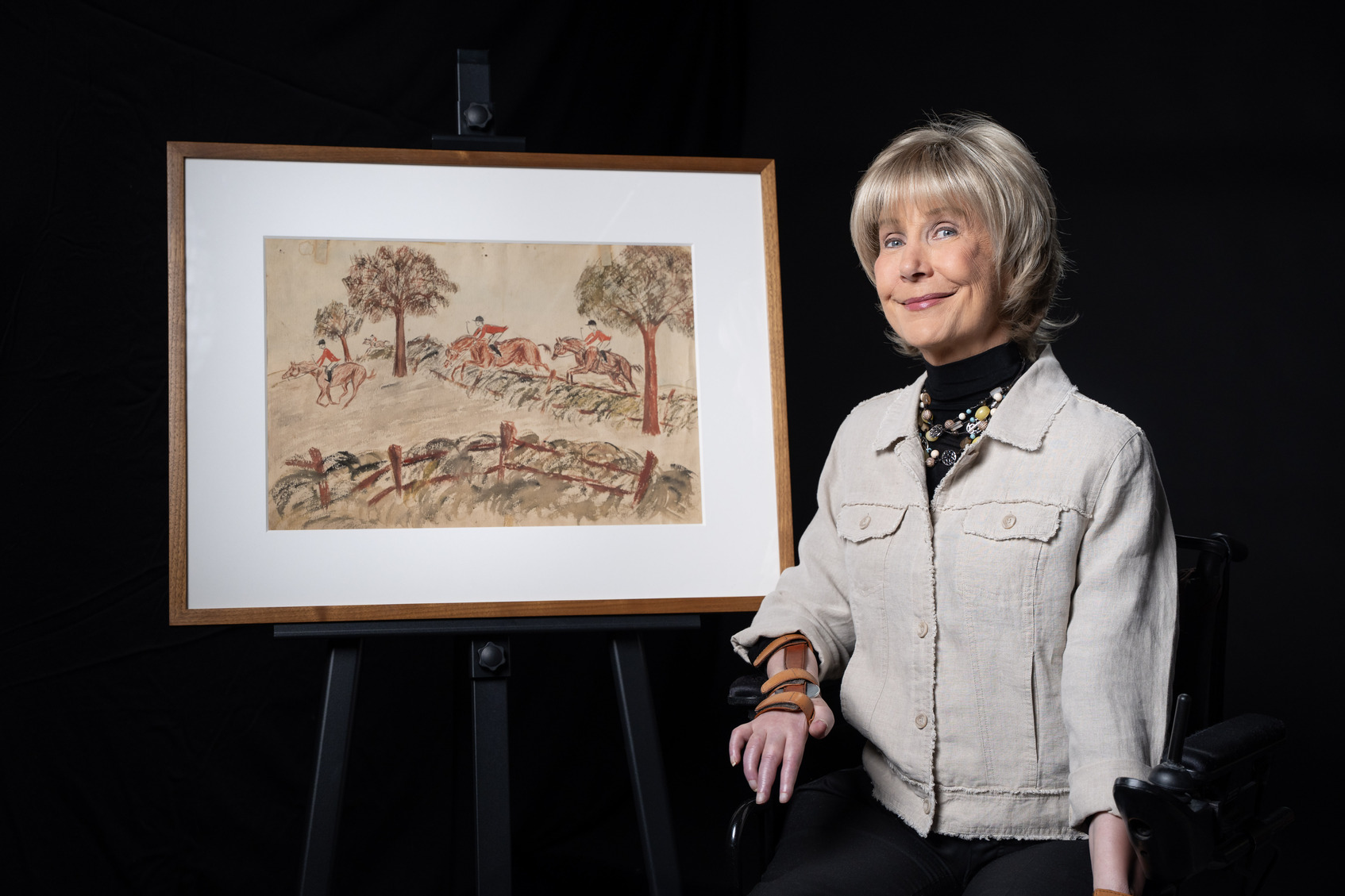 Joni Eareckson Tada next to her artwork titled "Steeplechase," which depicts three horses, each with a rider, jumping over hurdles.