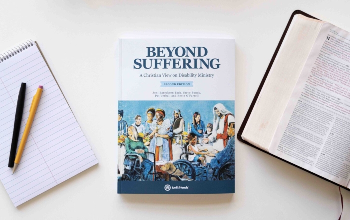 Book cover of Beyond Suffering: A Christian View on Disability Ministry, Second Edition, featuring the Luke 14 Banquet Mural where Jesus welcomes people with diverse disabilities. An open Bible is on the right, open to Luke 14, and a notepad with a pencil and pen is on the left.