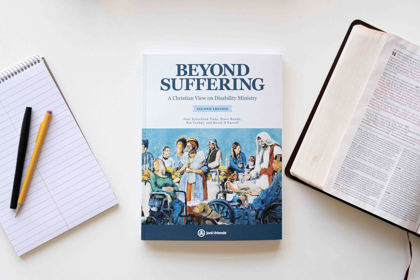 Book cover of Beyond Suffering: A Christian View on Disability Ministry, Second Edition, featuring the Luke 14 Banquet Mural where Jesus welcomes people with diverse disabilities. An open Bible is on the right, open to Luke 14, and a notepad with a pencil and pen is on the left.