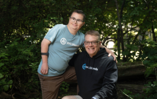 Paul, a young man with Down syndrome, standing beside Shawn, who is seated, both smiling and posing for the camera at Family Retreat.