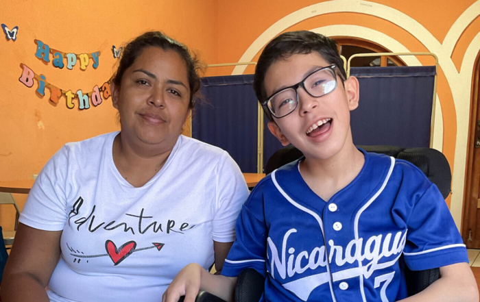 Zoomed-in image of Axel Rios, a smiling young boy in a wheelchair wearing glasses and a sports jersey, sitting on the right and holding his mother’s hand. His mother is seated beside him on the left. Behind them is a “Happy Birthday” banner, arched doorways, and a fan on a tiled floor.
