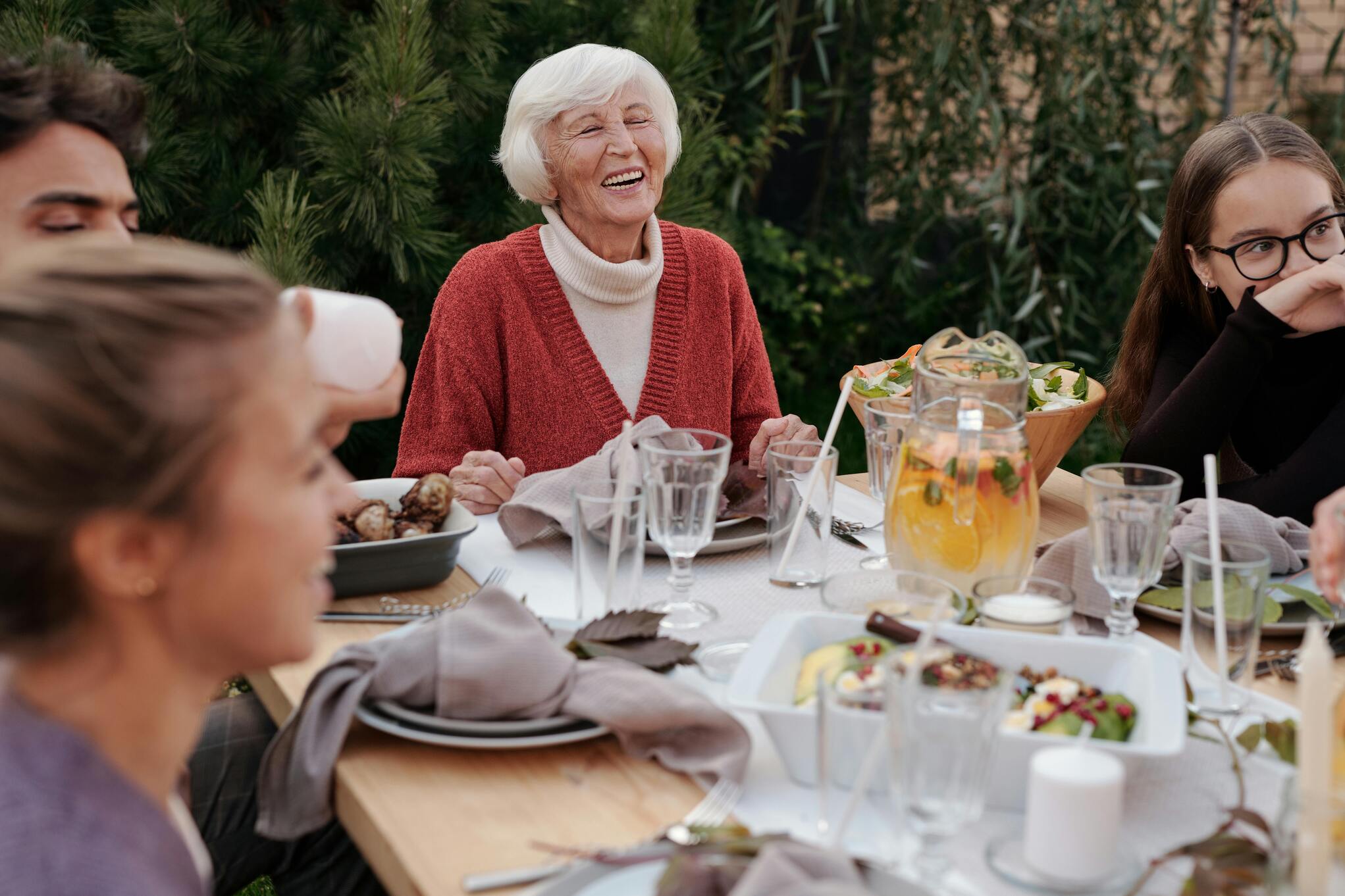 An elderly woman with a joyful, beautiful laugh is surrounded by family around a dinner table, sharing a warm, lively moment together.