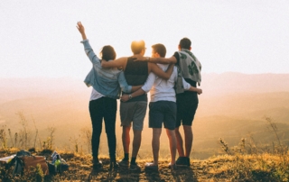 Four friends standing on top of a mountain, with their arms around each other's shoulders and backs, looking out at the scenic view together.