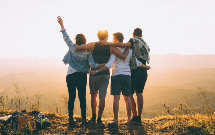 Four friends standing on top of a mountain, with their arms around each other's shoulders and backs, looking out at the scenic view together.