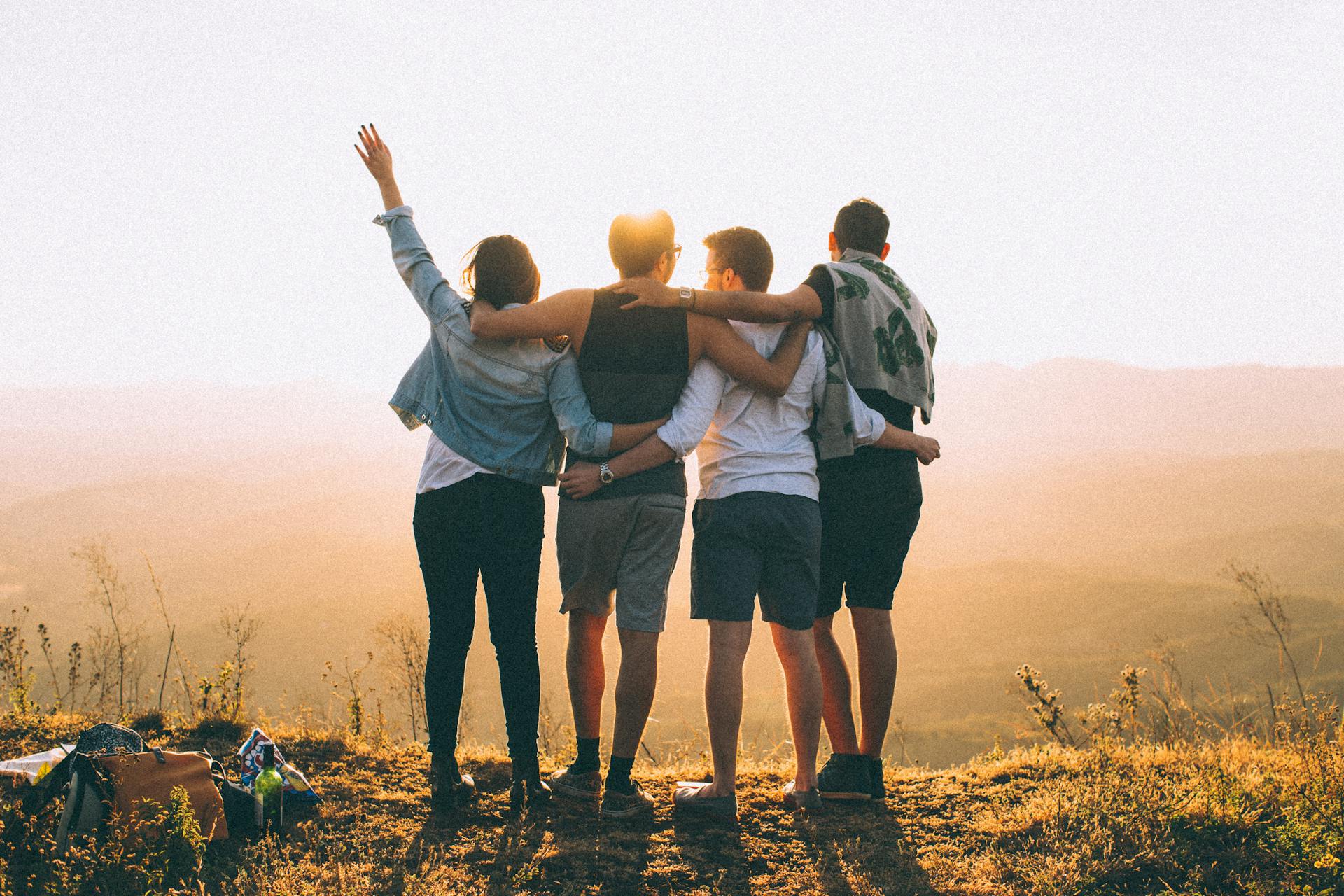 Four friends standing on top of a mountain, with their arms around each other's shoulders and backs, looking out at the scenic view together.